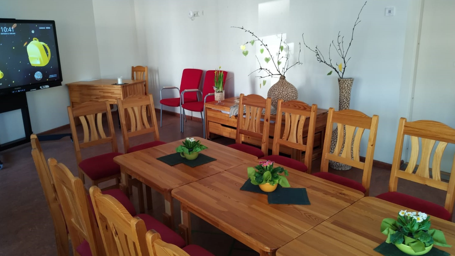 Rīgas 275. pirmsskolas izglītības iestādes “Austriņa” pasākuma telpa ar koka galdu, krēsliem un ekrānu