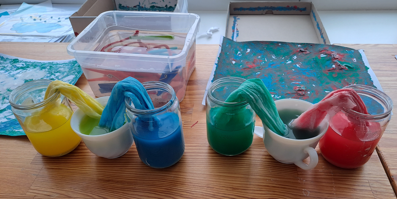 Dzeltena un zila, zaļa un sarkana krāsu šķīduma burkas savienotas pa pāriem ar  auduma strēmelēm tā, lai, krāsām sajaucoties, veidotos cita krāsa