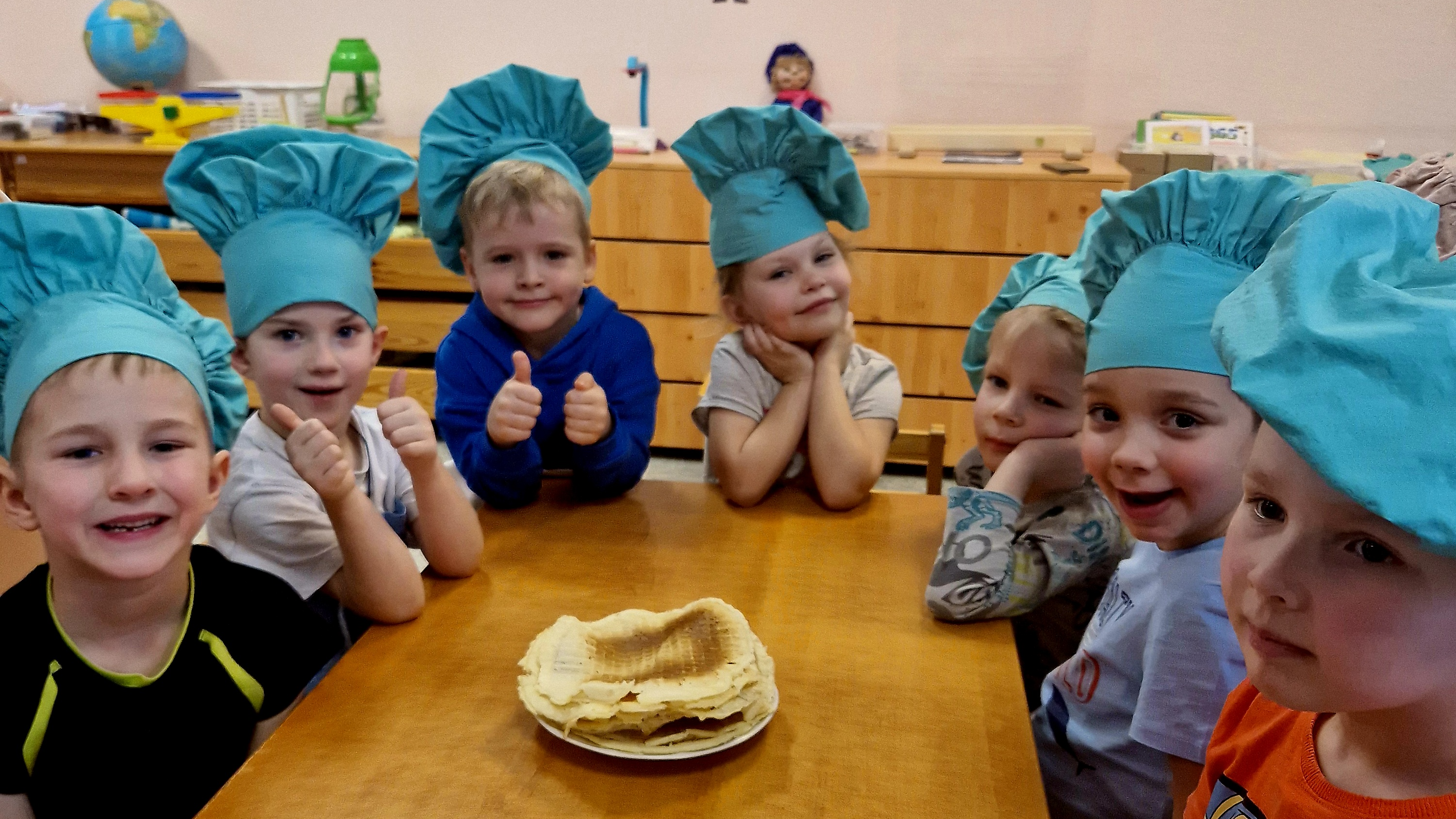 Bērnudārza grupiņas bērni apsēdušies ap galdu pavāru cepurēs pēc pīrādziņu cepšanas